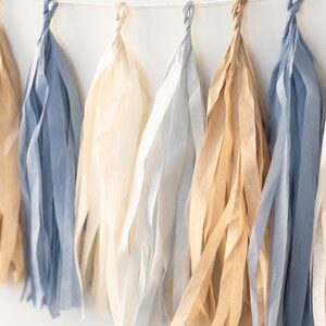Puede incluir: Una guirnalda de borlas de papel de seda en tonos beige, azul claro y marr&oacute;n. Las borlas est&aacute;n enhebradas con una cuerda blanca.