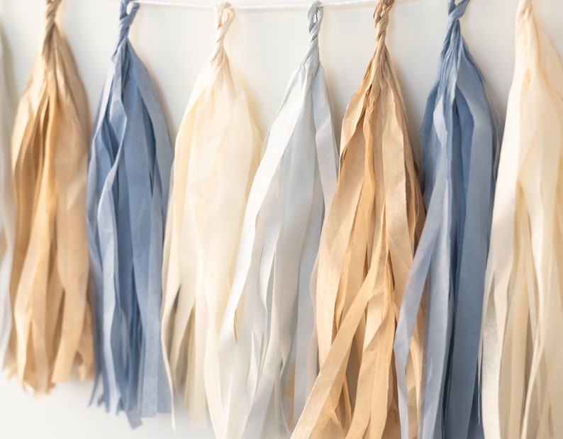 Puede incluir: Una guirnalda de borlas de papel de seda en tonos beige, azul claro y marr&oacute;n. Las borlas est&aacute;n enhebradas con una cuerda blanca.