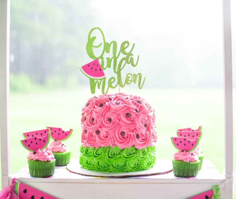 One in a Melon First Birthday  One in a Melon Cake Topper shown on a watermelon cake (pink icing cake with green on the bottom surrounded by small cupcakes with watermelon cupcake toppers)