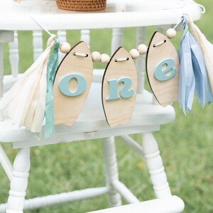 May include: A white high chair with a wooden surfboard banner that says "one" in blue letters. The banner is decorated with blue and white tissue paper tassels.
