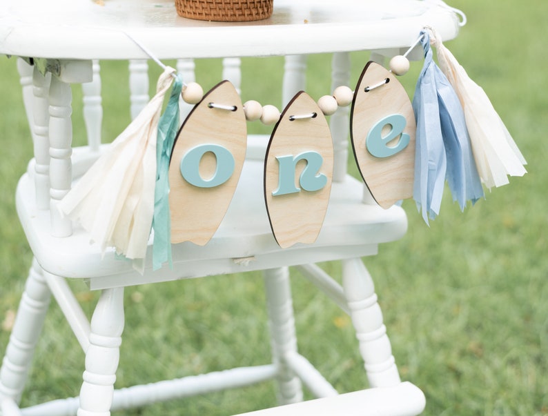 May include: A white high chair with a wooden surfboard banner that says "one" in blue letters. The banner is decorated with blue and white tissue paper tassels.