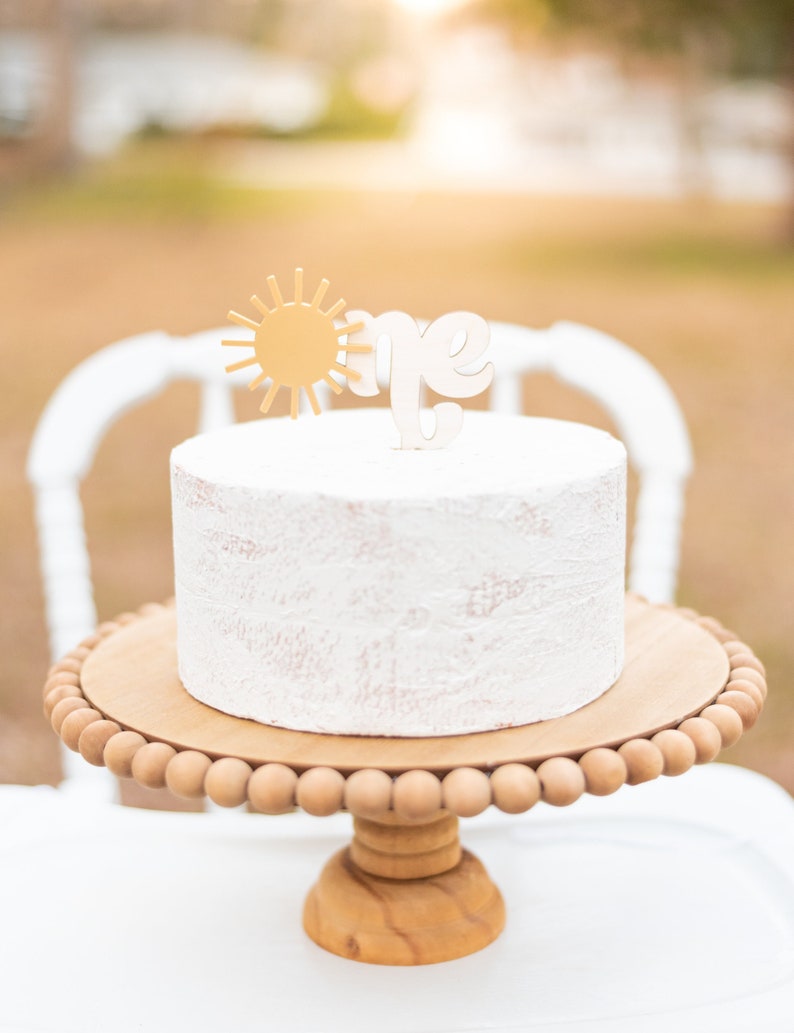Puede incluir: Un pastel blanco con un sol dorado y un adorno de "uno" blanco sobre un soporte para pasteles de madera con un borde de cuentas.