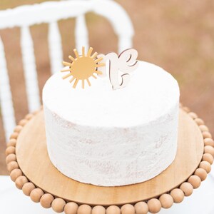 Puede incluir: Un pastel blanco glaseado con un sol dorado y un adorno de madera "uno" en un soporte de pastel de madera con un borde de cuentas.