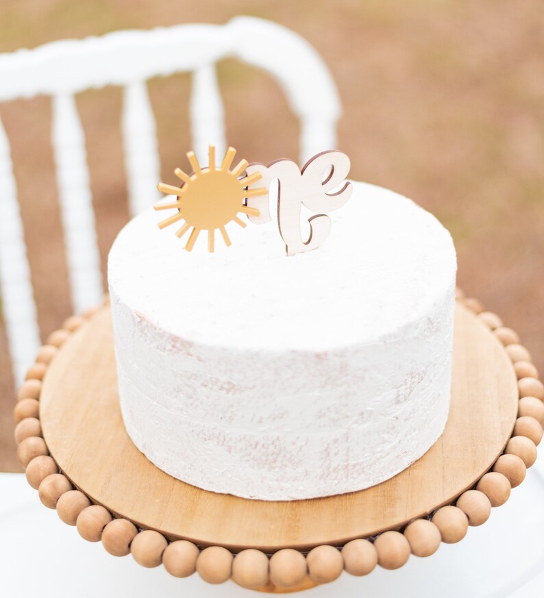 Puede incluir: Un pastel blanco glaseado con un sol dorado y un adorno de madera "uno" en un soporte de pastel de madera con un borde de cuentas.