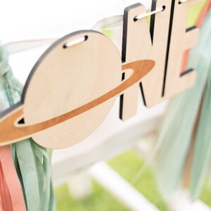 May include: Wooden banner with the word "ONE" in large letters, decorated with a planet and ring design. The banner is strung with white twine and has light blue and pink streamers attached.