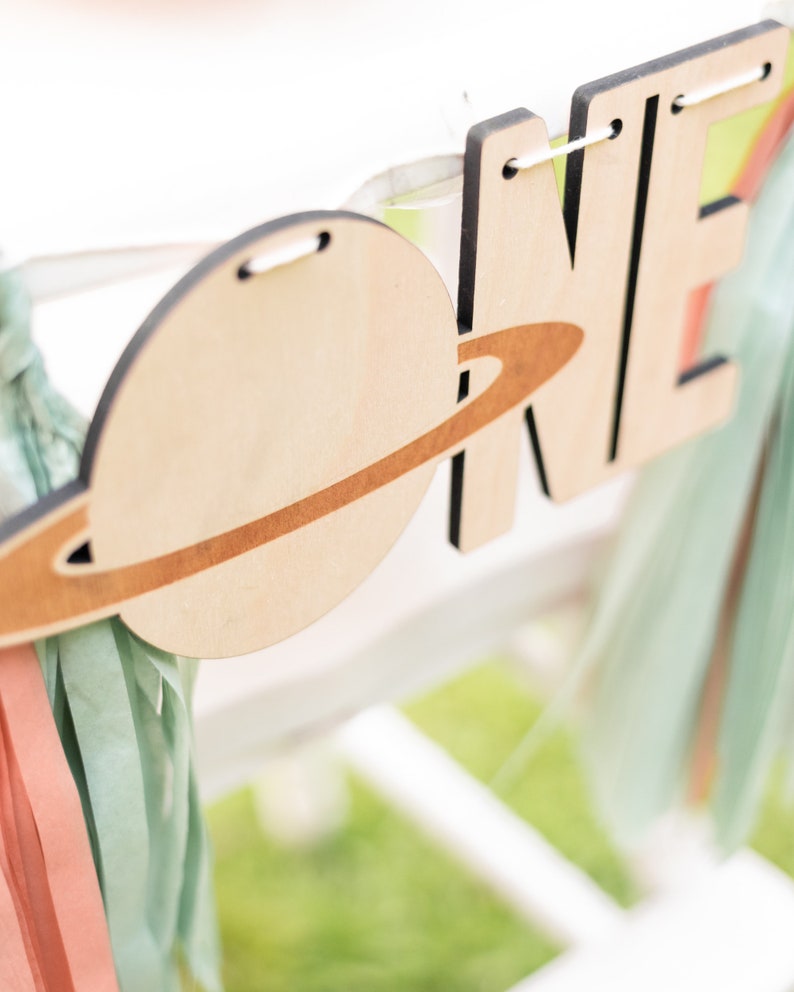 May include: Wooden banner with the word "ONE" in large letters, decorated with a planet and ring design. The banner is strung with white twine and has light blue and pink streamers attached.