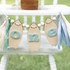 May include: A wooden "one" banner with blue letters and wooden surfboard shapes, strung with wooden beads and white, blue, and cream tissue paper tassels.