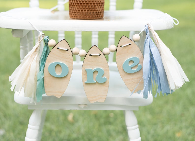 May include: A wooden "one" banner with blue letters and wooden surfboard shapes, strung with wooden beads and white, blue, and cream tissue paper tassels.