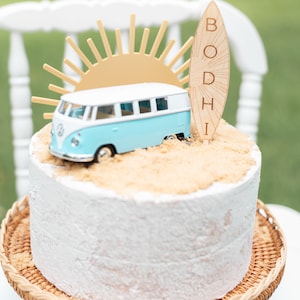 May include: A white cake with blue and white toy van, a yellow sun, and a wooden surfboard with the name "BODHI" on it. The cake is on a wicker cake stand.