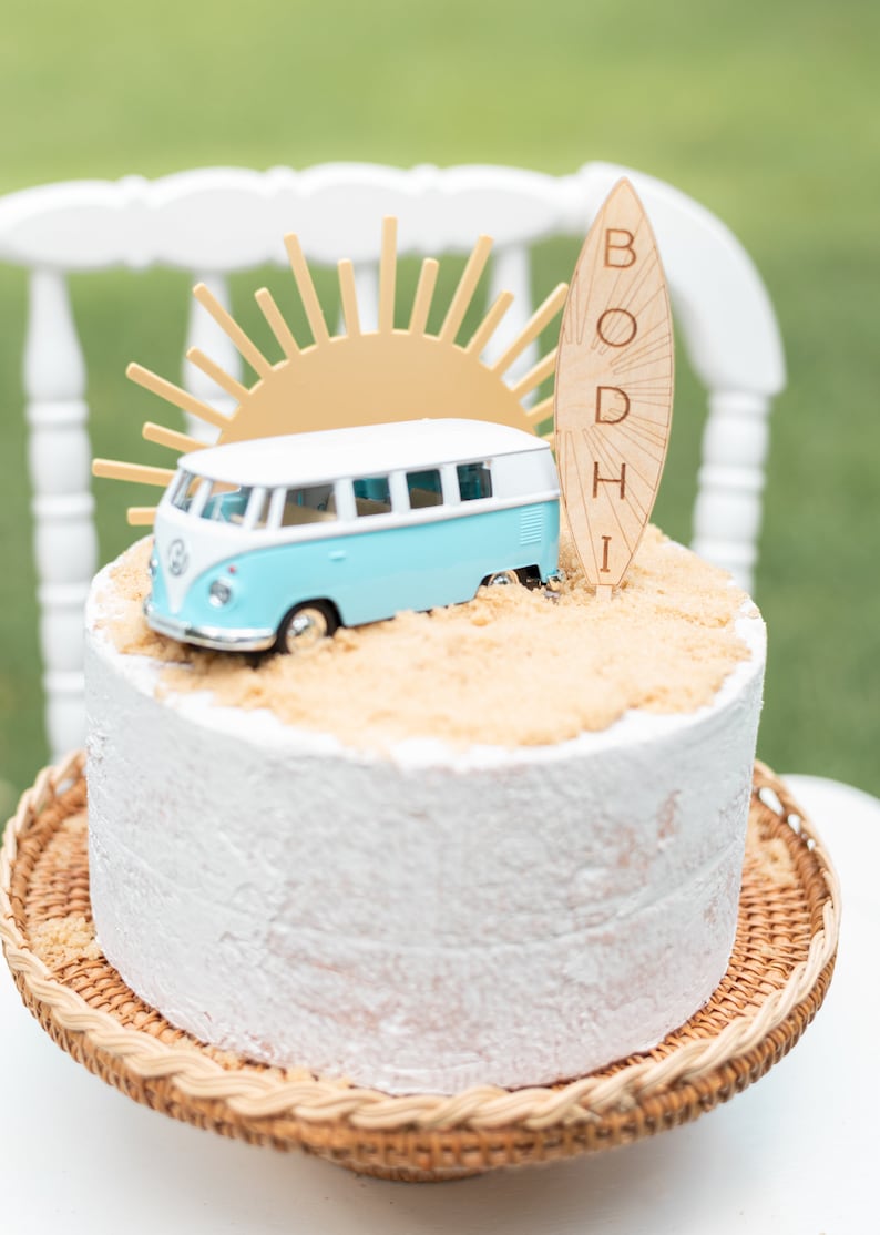 May include: A white cake with blue and white toy van, a yellow sun, and a wooden surfboard with the name "BODHI" on it. The cake is on a wicker cake stand.