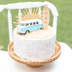 May include: A white cake with a blue and white toy van, a gold sun, and a wooden surfboard with the name "BODHI" on it. The cake is on a brown wicker cake stand.