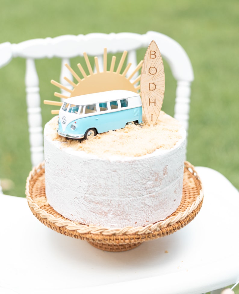 May include: A white cake with a blue and white toy van, a gold sun, and a wooden surfboard with the name "BODHI" on it. The cake is on a brown wicker cake stand.