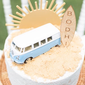 May include: A white and blue toy van sits on a cake decorated with brown sugar to look like sand. A wooden surfboard with the name "BODHI" is leaning against the van. A yellow paper sun sits behind the van.