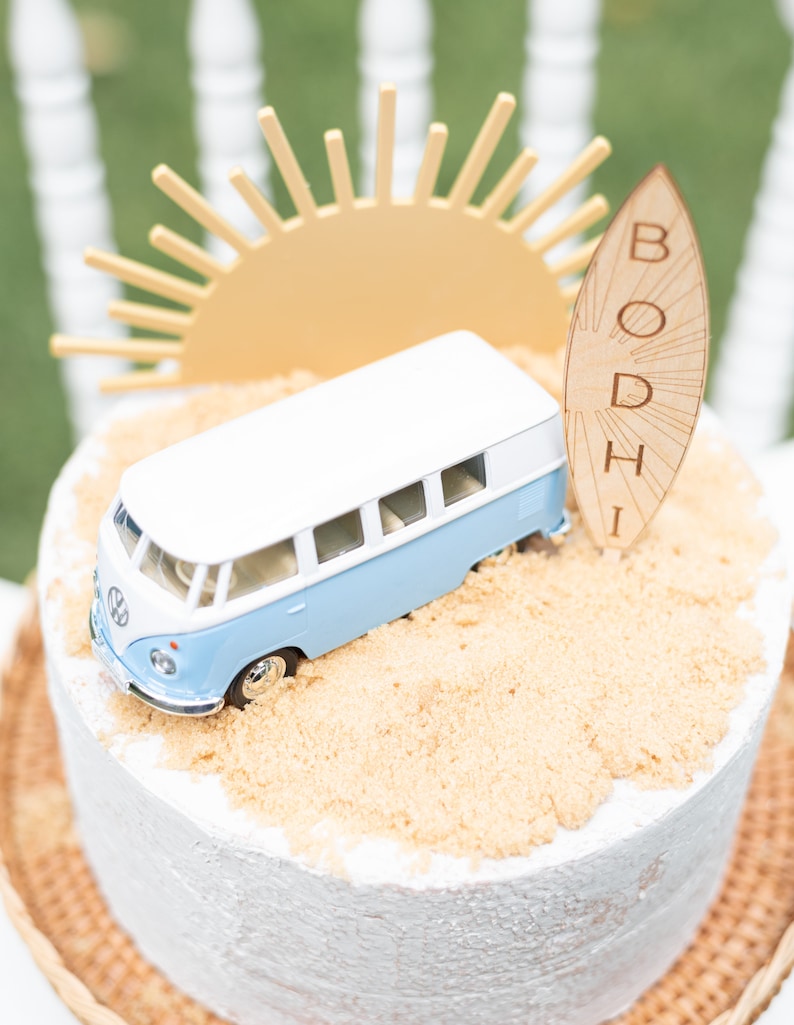 May include: A white and blue toy van sits on a cake decorated with brown sugar to look like sand. A wooden surfboard with the name "BODHI" is leaning against the van. A yellow paper sun sits behind the van.