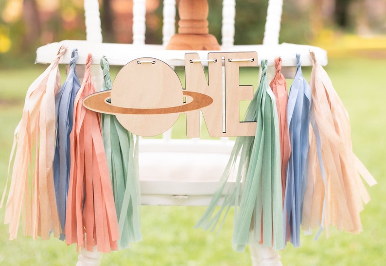 May include: A wooden "ONE" banner with a planet and rings decoration, hanging from a white chair with pastel colored tissue paper tassels.