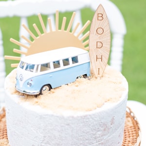 May include: A white cake decorated with a blue and white toy van, a wooden surfboard with the name "BODHI" written on it, and a gold sun.