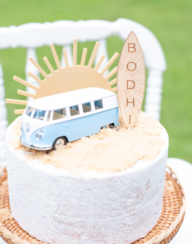 May include: A white cake decorated with a blue and white toy van, a wooden surfboard with the name "BODHI" written on it, and a gold sun.