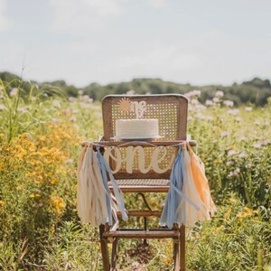 Puede incluir: Una silla alta de madera con un pastel blanco en la parte superior y una pancarta que dice "one" en frente. La silla est&aacute; decorada con borlas azules y melocot&oacute;n. La silla est&aacute; en un campo de flores amarillas.