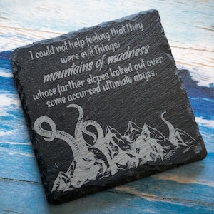 May include: Square black slate coaster with engraved text and a mountain and tentacle design. The text reads: "I could not help feeling that they were evil things; mountains of madness whose farther slopes looked out over some accursed ultimate abyss."