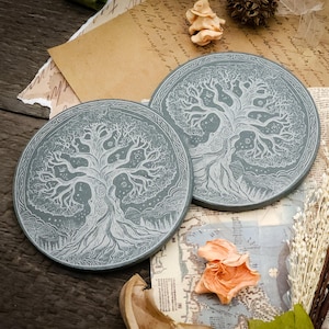 Pode incluir: Duas bases redondas cinzentas com um design branco de árvore da vida. As bases são feitas de pedra e têm um padrão gravado detalhado. A imagem é estilizada com flores secas e papel vintage.