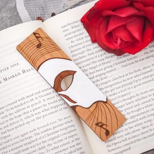 May include: A wooden bookmark with a white mask design and musical notes. The bookmark is laying on a book with the text "Chapter Nine At The Masked Ball".