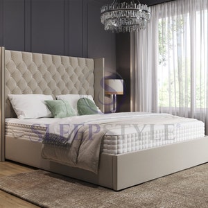 May include: A beige upholstered bed frame with a tall headboard featuring a tufted design. The bed is made up with white bedding and green pillows. The bed is in a bedroom with a white rug, a nightstand, and a chandelier.