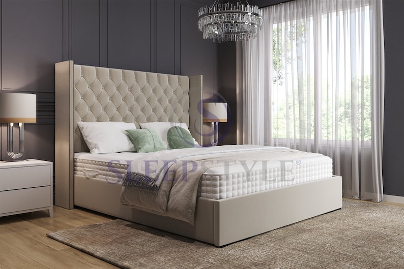 May include: A beige upholstered bed frame with a tall headboard featuring a tufted design. The bed is made up with white bedding and green pillows. The bed is in a bedroom with a white rug, a nightstand, and a chandelier.