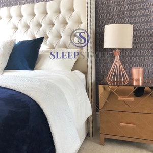 May include: A white tufted headboard with a decorative nailhead trim, a white and blue throw blanket, and a white pillow with the text "SLEEPSTYLE" on it. A rose gold bedside table with a mirrored surface and a rose gold lamp with a white shade are on the right side of the image.