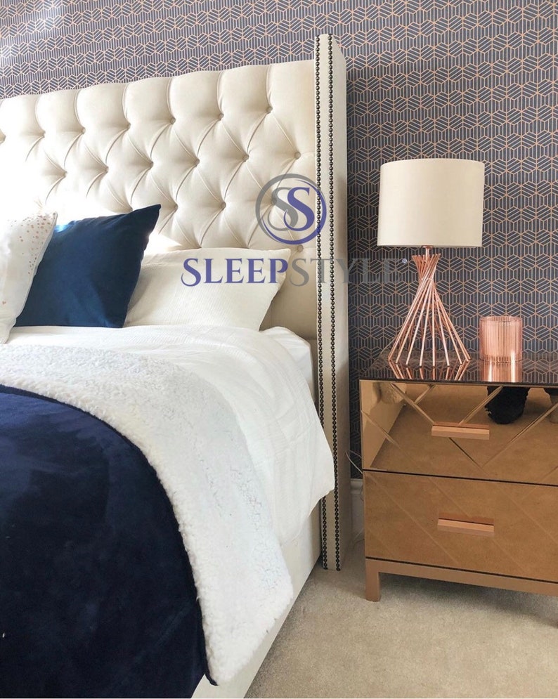 May include: A white tufted headboard with a decorative nailhead trim, a white and blue throw blanket, and a white pillow with the text "SLEEPSTYLE" on it. A rose gold bedside table with a mirrored surface and a rose gold lamp with a white shade are on the right side of the image.