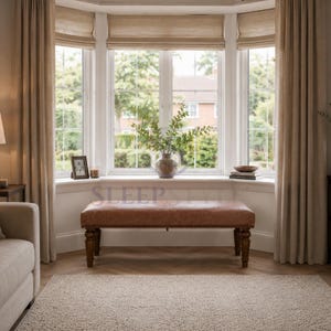 May include: A rectangular brown leather bench with a tufted top and dark wooden turned legs positioned in front of a large bay window. The bench features nailhead trim along the base and sits on a light-coloured rug in a room with beige curtains.