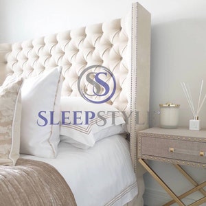 May include: A white tufted headboard with a tall, upholstered wingback design. The headboard is accented with silver nailhead trim. The bed is made with white bedding and a beige throw blanket. A small, beige bedside table with a gold metal frame sits next to the bed.