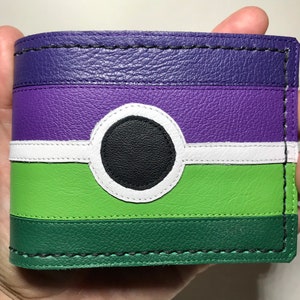 May include: A bi-fold wallet with a colorful design. The top section is purple, followed by a white stripe, a green section, and a dark green base. A black circle is centered on the front, outlined in white. The wallet is held in a hand.