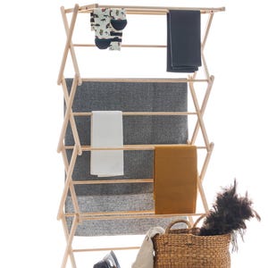 Könnte beinhalten: Ein Wäscheständer aus Holz mit verschiedenen Textilien, darunter eine graue Decke, ein weißes Handtuch und eine senffarbene Decke. Ein Weidenkorb, ein Bügeleisen und ein Staubwedel sind ebenfalls vorhanden. Socken mit einem Automuster hängen auf dem Ständer.