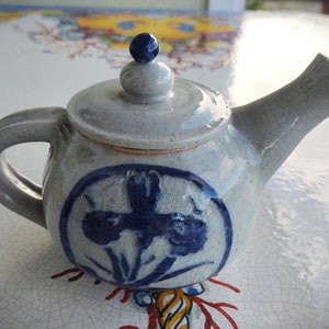 May include: A small, gray ceramic teapot with a blue floral design. The teapot has a round body and a spout on the side. The lid has a small knob on top.