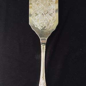 May include: Silver-colored serving spatula with an ornate, engraved floral design on the blade. The handle is long and slender, with a decorative pattern at the base. The spatula is likely for serving cakes or other desserts.