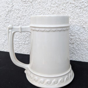Puede incluir: Una jarra de cerveza de cerámica blanca con asa y detalles decorativos alrededor de la parte superior e inferior.