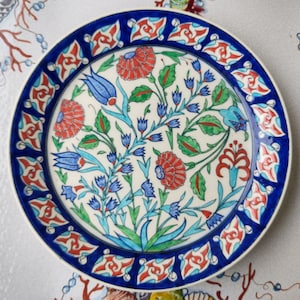 Puede incluir: Un plato de cerámica blanco con un borde azul y rojo. El centro del plato presenta un diseño floral con flores azules, rojas y verdes.