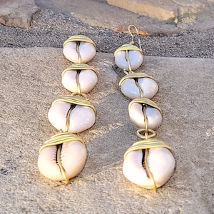 May include: A pair of long, gold-toned earrings with white cowrie shells. Each earring has four shells strung together with wire.