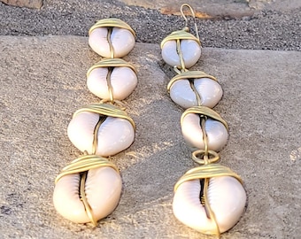 May include: A pair of long, gold-toned earrings featuring white cowrie shells. Each earring has four shells strung together with wire.