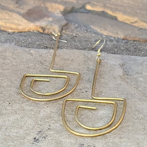 May include: A pair of gold geometric earrings with a curved, layered design. The earrings are made of a single piece of wire that is bent into a unique shape.