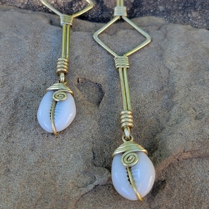 May include: A pair of long, gold-colored earrings. Each earring features a diamond-shaped top, a long vertical bar, and a white, oval-shaped shell pendant. The shells are wrapped in gold wire, and the earrings have a hook closure.