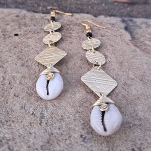 May include: Gold-toned dangle earrings featuring a series of geometric shapes: circles, a diamond, and a cowrie shell. The diamond shape has etched lines. The earrings have a hook closure and black bead accents.