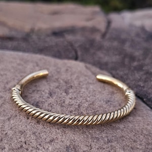 May include: A gold-colored cuff bracelet with a twisted rope design. The open-ended bracelet is resting on a gray stone surface. The bracelet is a simple, yet elegant piece of jewelry.