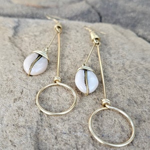 May include: A pair of gold earrings with a cowrie shell charm hanging from a long, thin wire. The earrings have a simple, minimalist design and are perfect for everyday wear.