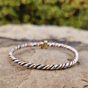 May include: A silver, gold, and copper braided bangle bracelet. The bracelet is twisted and has a smooth finish.