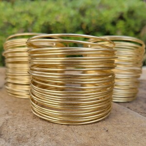 May include: Three gold-colored wire bracelets. Each bracelet is made of numerous thin, tightly wound wires, creating a stacked effect. The bracelets are displayed on a stone surface with a blurred green background.