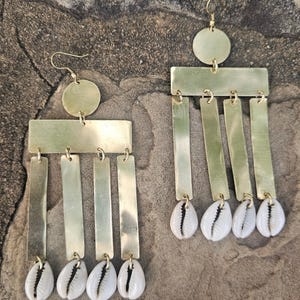 May include: A pair of brass earrings with a circular top, a rectangular middle section, and dangling rectangular bars. Each bar is adorned with a white cowrie shell. The earrings are designed for pierced ears.