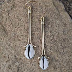 May include: A pair of long, gold-toned earrings featuring a cowrie shell design. The earrings have a slender, elongated shape with a hook closure. The cowrie shells are white with a black stripe down the center.