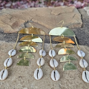May include: Gold-toned, layered earrings featuring a series of half-moon shapes and dangling chains. Each earring has three strands of white cowrie shells. The earrings are designed with a hook closure.