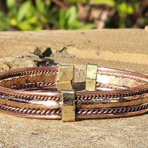 May include: A copper and brass cuff bracelet with a geometric design. The bracelet features alternating bands of copper and brass, with a central band of woven copper. The clasp is made of gold-colored metal.